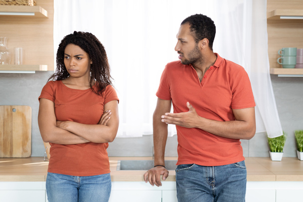 Sad angry young black husband swears at wife, offended sad woman in red t-shirt ignores husband after quarrel in kitchen interior. Stress and domestic violence, scandal at home, relationship problems - Photo, Image