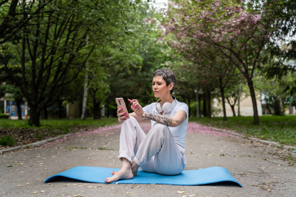 jeden Dojrzały kaukaski kobieta senior kobieta siedzieć odkryty na jogi mat używać telefon komórkowy dla online szkolenia lub przewodnik medytacja praktyka self care emocjonalny równowaga koncepcja kopia przestrzeń - Zdjęcie, obraz