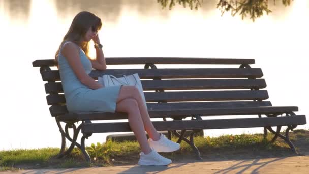Young woman in summer dress sitting relaxed on lake side bench on warm evening. Wellness resting from everyday rush concept. - Footage, Video