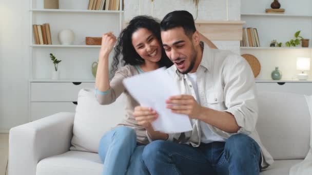 Euphoric multiracial ethnic couple sit on sofa woman and man happy reading document paper letter with amazing winner victory offer news bank loan approval profitable job opportunity prize celebrating - Footage, Video