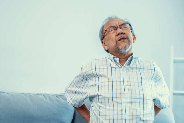 Ένας αγωνιώδης ηλικιωμένος που χρειάζεται βοήθεια ενώ κάθεται στον καναπέ του στο σπίτι, και υποφέρει από πόνο στην πλάτη. Ανώτερη φροντίδα, γηροκομείο συνταξιούχων, επιδείνωση της υγείας των ηλικιωμένων. - Φωτογραφία, εικόνα
