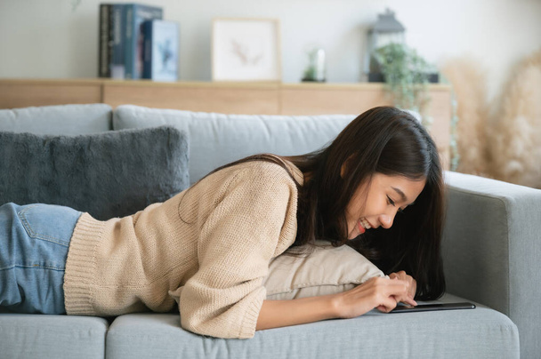 Feliz joven asiática mujer relajarse en cómodo sofá en casa mensajes de texto de mensajería en el teléfono inteligente, chica sonriente utilizar el teléfono celular, navegar por Internet inalámbrico en gadget, compras en línea desde casa - Foto, imagen
