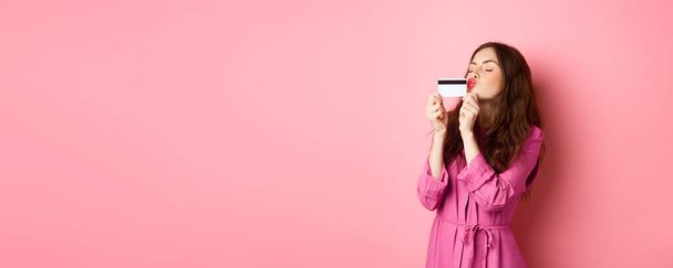 Imagem da jovem mulher bonita beijando seu cartão de crédito, indo às compras, desperdiçando dinheiro nas lojas, de pé contra fundo rosa. Espaço de cópia - Foto, Imagem