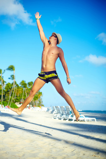 Happy handsome muscled man in sunhat on beach jumping behind blue sky behind blue sky - Фото, зображення