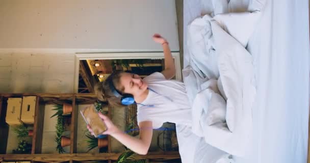 Imágenes de 4k de una niña de 10 años sentada en una cama con auriculares y escuchando música a través de la aplicación para teléfonos inteligentes. Vídeo vertical. - Metraje, vídeo