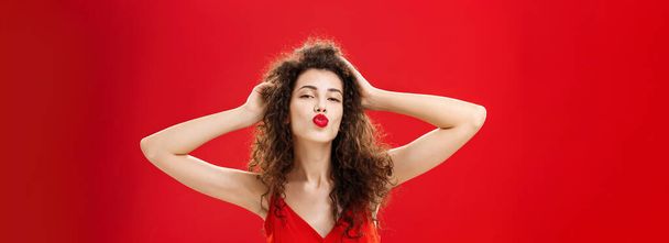 Waist-up shot of confident flirty and elegant young rich woman. with curly hairstyle in red lipstic and evening dress touching hair folding lips in mwah giving kiss to admirer feeling self-assured. - Photo, Image