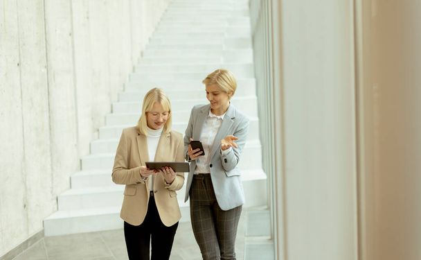 Dvě obchodní ženy kráčející s notebookem a mobilním telefonem na chodbě kanceláře - Fotografie, Obrázek