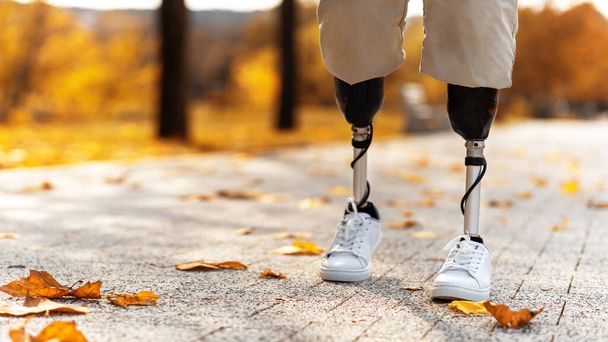 Veduta di un uomo che cammina con gambe protesiche e scarpe da ginnastica bianche. Foglie gialle cadute a terra - Foto, immagini