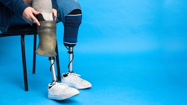 Protez bacaklı ve beyaz spor ayakkabılı bir adam görüntüsü. Bir sandalyede otururken protezi çıkarmak, mavi arkaplan - Fotoğraf, Görsel