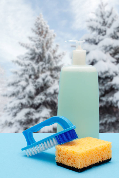 Bouteille de liquide vaisselle, brosse et éponge avec paysage hivernal sur le fond. Concept de lavage et de nettoyage. - Photo, image