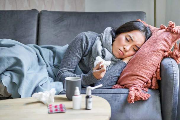 Mulher coreana triste doente deitada no sofá, sentindo-se mal, resfriado, gripe e temperatura, parecendo chateada, tomando medicação. - Foto, Imagem
