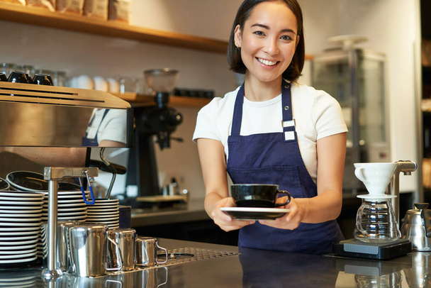 Portret van een glimlachende jonge vrouw die klantenservice verleent in een café, een kop koffie vasthoudt, een bestelling geeft aan de klant, een schortenuniform draagt. - Foto, afbeelding