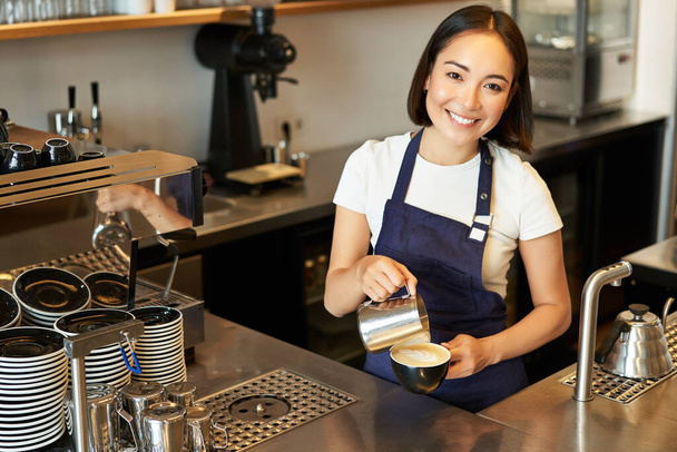 Mosolygó ázsiai lány barista, kávézó személyzet öntés gőzölt tej kávé, készítsen cappuccino latte art, álló kék kötény a pult mögött. - Fotó, kép