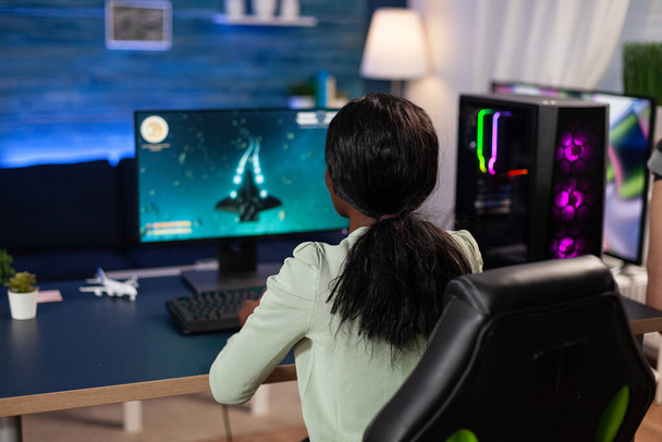 Femme joueuse professionnelle assise au bureau de jeu en jouant aux jeux vidéo de tir de l'espace pour le championnat en ligne. Joueur concentré utilisant un équipement informatique RVB professionnel en home studio. Jeux de sport électronique - Photo, image