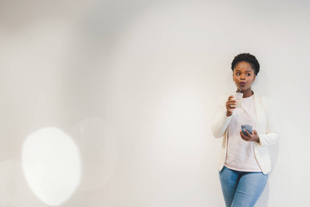 Bild einer fröhlichen erwachsenen afrikanischen Frau, die ein Glas Kaffee trinkt und ihr Handy benutzt, während sie sich an die weiße Wand mit Kopierraum lehnt. Leere Räume. - Foto, Bild