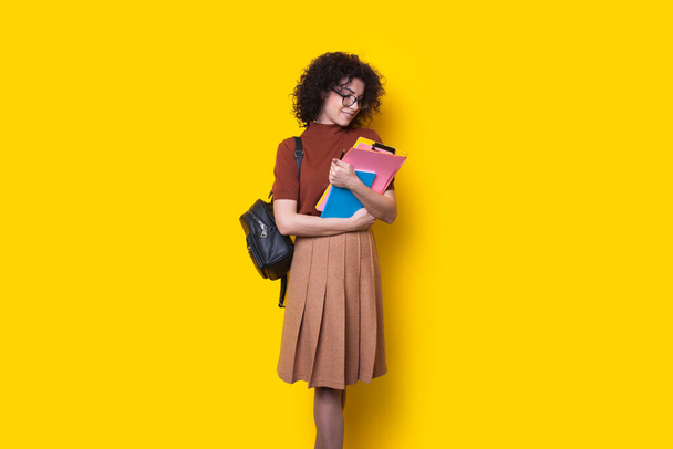 Σγουρομάλλη μαθήτρια με σακίδιο που κρατάει κάποια έγγραφα και βιβλία απομονωμένα σε κίτρινο φόντο. Εκπαίδευση στο πανεπιστήμιο γυμνασίου - Φωτογραφία, εικόνα