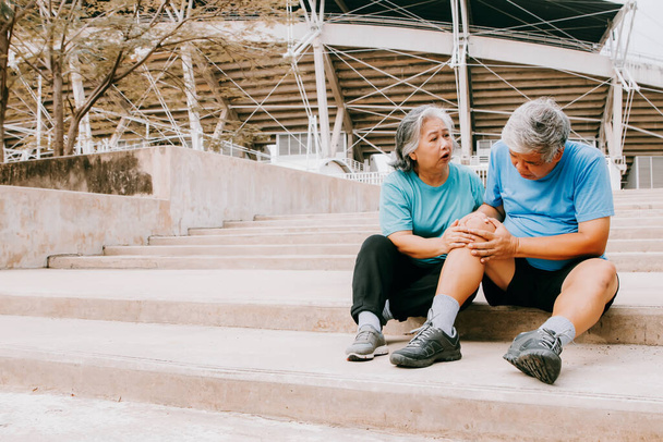 Starszy mężczyzna z bólem w prawym kolanie podczas joggingu po twardych betonowych schodach stadionu sportowego ma nasilony ból w jego rannym kolanie, pod opieką żony: Starsza koncepcja ubezpieczenia zdrowotnego - Zdjęcie, obraz
