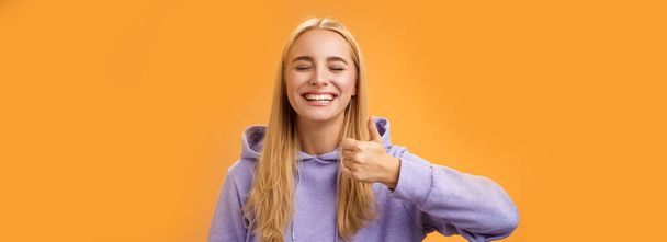 Szórakoztató vidám optimista fiatal izgatott szőke európai nő modell kapucnis mosoly tágan csukott szemmel szórakozás show jóváhagyás gesztus hüvelykujj-up, mint félelmetes kirándulás nyaralás ötlet, narancs háttér. - Fotó, kép