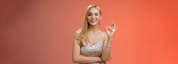 Sassy blond young woman in stylish luxury silver dress winking take things under control show okay no problem gesture make ok sign, like awesome party praise excellent effort, red background. - Photo, Image