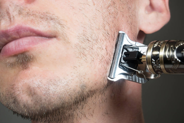 Elektrikli metal ustura ya da kuru tıraş makinesiyle sakal kesiyor. Kesmek için budayıcı. Erkek yüzünde kısa, seyrek bir sakal. Saç uzama problemleri. Sakalında tıraş edilmemiş kıllar var.. - Fotoğraf, Görsel