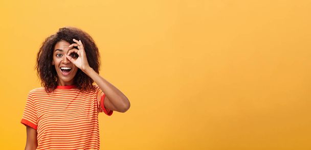 Creative and playful dreamy dark-skinned adult girl with curly hairstyle in striped t-shirt showing circle over eye or okay gesture smiling broadly ready take part in adventures near orange wall - Photo, Image