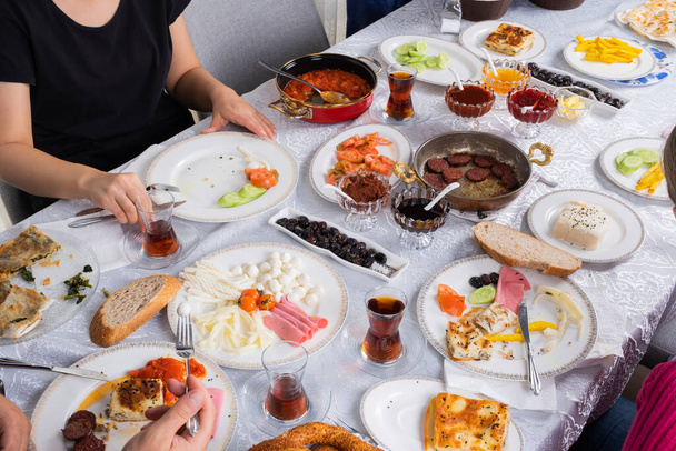 Traditional modern Turkish family breakfast table, over shoulder view. Classic home breakfast. Cheese, borek, sucuk, cheese, tea in glass, fried potatoes, bread, black olive, simit, honey on the table - Foto, immagini