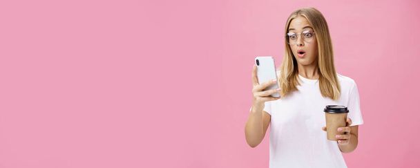 Γυναίκα πίνοντας καφέ είναι συγκλονισμένοι από έλαβε μήνυμα αντιδρώντας σε εκπληκτική είδηση αναδίπλωση χείλη λαχανιάζοντας αναζητούν έκπληκτος και εντυπωσιασμένος σε οθόνη smartphone κρατώντας χάρτινο κύπελλο, θέτοντας πάνω από το ροζ τοίχο - Φωτογραφία, εικόνα