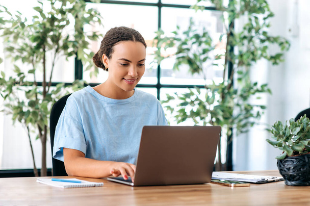 Positiivinen kaunis sekarotuinen liiketoiminnan nainen istuu pöydässä modernissa toimistossa, työskentelee kannettavassa tietokoneessa projektissa, selailee tietoja Internetissä, vastaa sähköpostiin, hymyilee - Valokuva, kuva