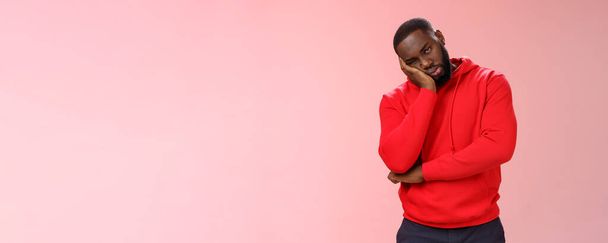 Bored young 25s black guy in red hoodie facepalm lean head hand look indifferent sleepy attend boring meeting fall asleep carelessly listening uninteresting story, pink background. Copy space - Photo, Image