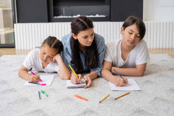 aziatische kids tekening op papier in de buurt moeder liggend op tapijt thuis  - Foto, afbeelding