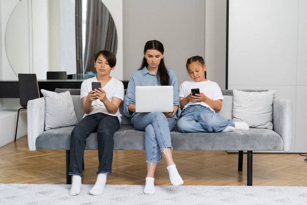 Woman using laptop near asian children with smartphones at home  - Foto, Bild