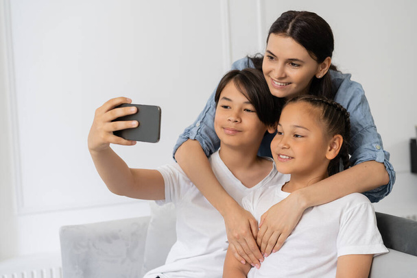 Mère souriante étreignant les enfants asiatiques tout en fils prenant selfie à la maison  - Photo, image