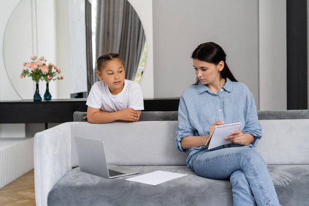 Asian kid looking at mom holding notebook near laptop at home  - Photo, Image