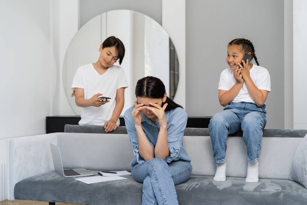 Tired mother sitting near laptop and asian kids using smartphones at home  - Valokuva, kuva