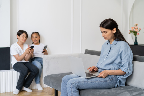 Mother using laptop near blurred asian children with cellphones at home  - Photo, Image