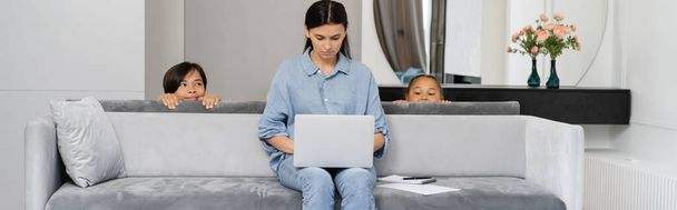 Sly asian kids looking at mom using laptop on couch at home, banner  - Фото, зображення