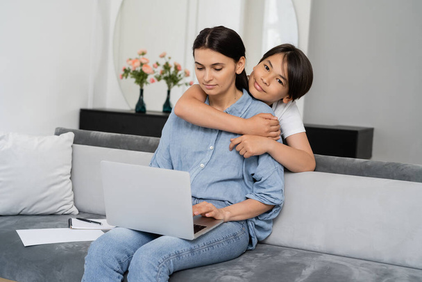 Lächelnd asiatisches Kind umarmt Mutter mit Laptop im Wohnzimmer  - Foto, Bild
