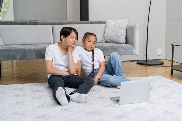 Asian siblings watching cartoons on laptop in living room  - Фото, изображение