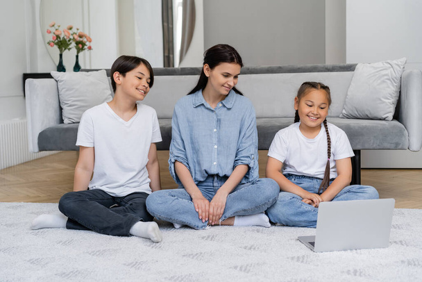 sonriente madre viendo película con asiático niños en portátil en casa  - Foto, imagen