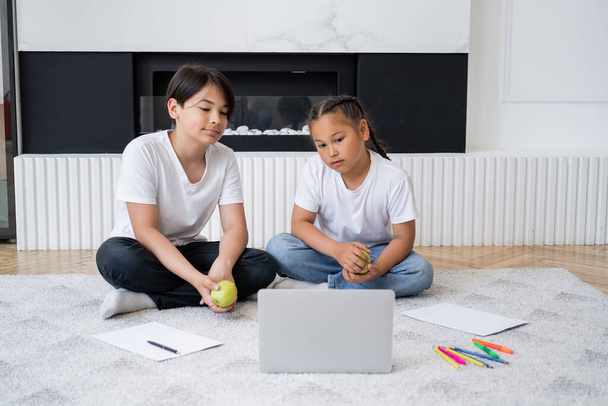 Asyalı çocuklar ellerinde elmalarla dizüstü bilgisayarlarında film izliyorlar. Evlerinde kağıt ve renkli kalemler.  - Fotoğraf, Görsel