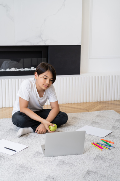 Preteen asiatique garçon tenant pomme près ordinateur portable et crayons de couleur à la maison  - Photo, image