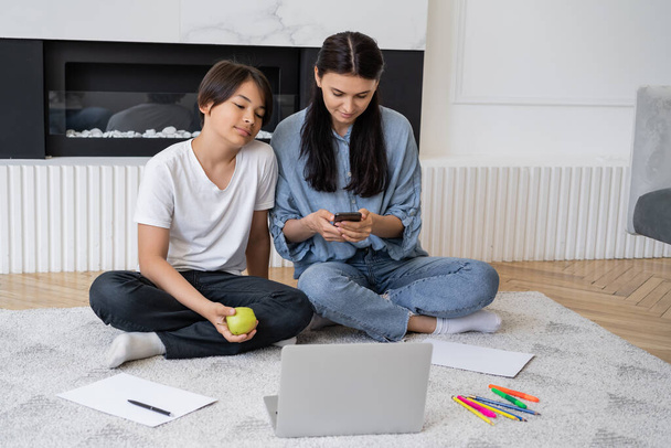 Frau benutzt Smartphone in Nähe asiatischen Sohnes mit Apfel Blick auf Laptop im Wohnzimmer  - Foto, Bild