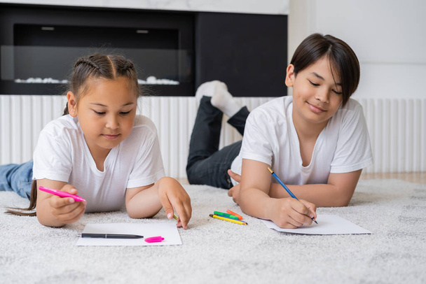 Smiling asian siblings drawing on papers on floor at home  - Photo, Image