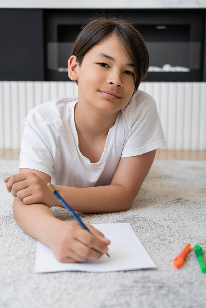 Preteen asiatique garçon regarder caméra tout en dessinant sur papier à la maison  - Photo, image