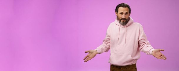 Bothered confused mature adult bearded man grey hair in pink hoodie arguing look pissed offended shrugging spread hands sideways dismay look clueless camera asking why, purple background. - Photo, Image