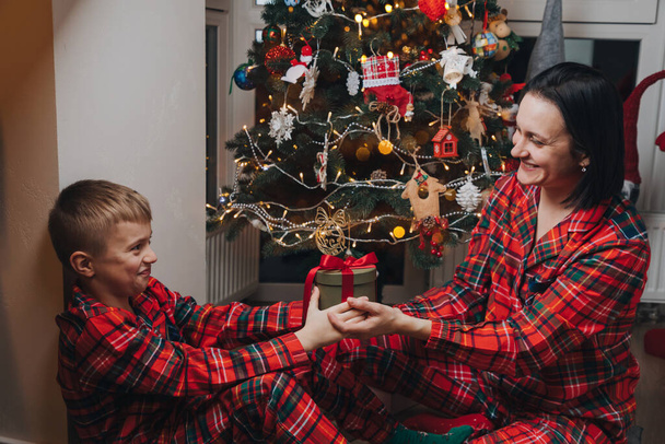 Mutter und Sohn sitzen im roten Schlafanzug neben dem Weihnachtsbaum und tauschen Geschenke aus. Frontansicht - Foto, Bild