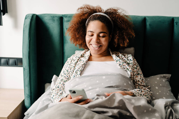 Молодая африканская американка улыбается и пользуется сотовым телефоном, лежа дома в постели - Фото, изображение