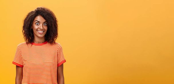 tiro en la cintura de la mujer afroamericana encantadora sorprendida y emocionada con el peinado rizado haciendo estallar los ojos de la emoción y la alegría sonriendo ampliamente sorprendido por el gran regalo sobre fondo naranja - Foto, imagen