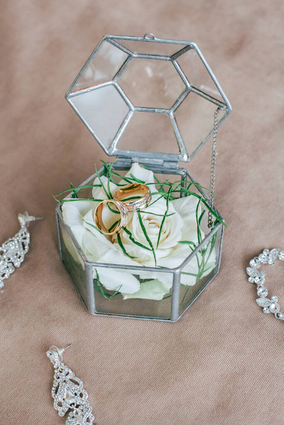 Декоративная стеклянная подарочная коробка для свадьбы, на бежевом фоне. Внутри золотые обручальные кольца для невесты и жениха, а также белая роза и зеленые листья. - Фото, изображение