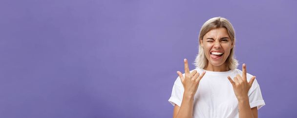 Gonna rock. Portrait of joyful happy good-looking stylish woman with blond medium haircut winking smiling and sticking out tongue while showing rock-n-roll gesture with both hands over purple wall. - Photo, Image